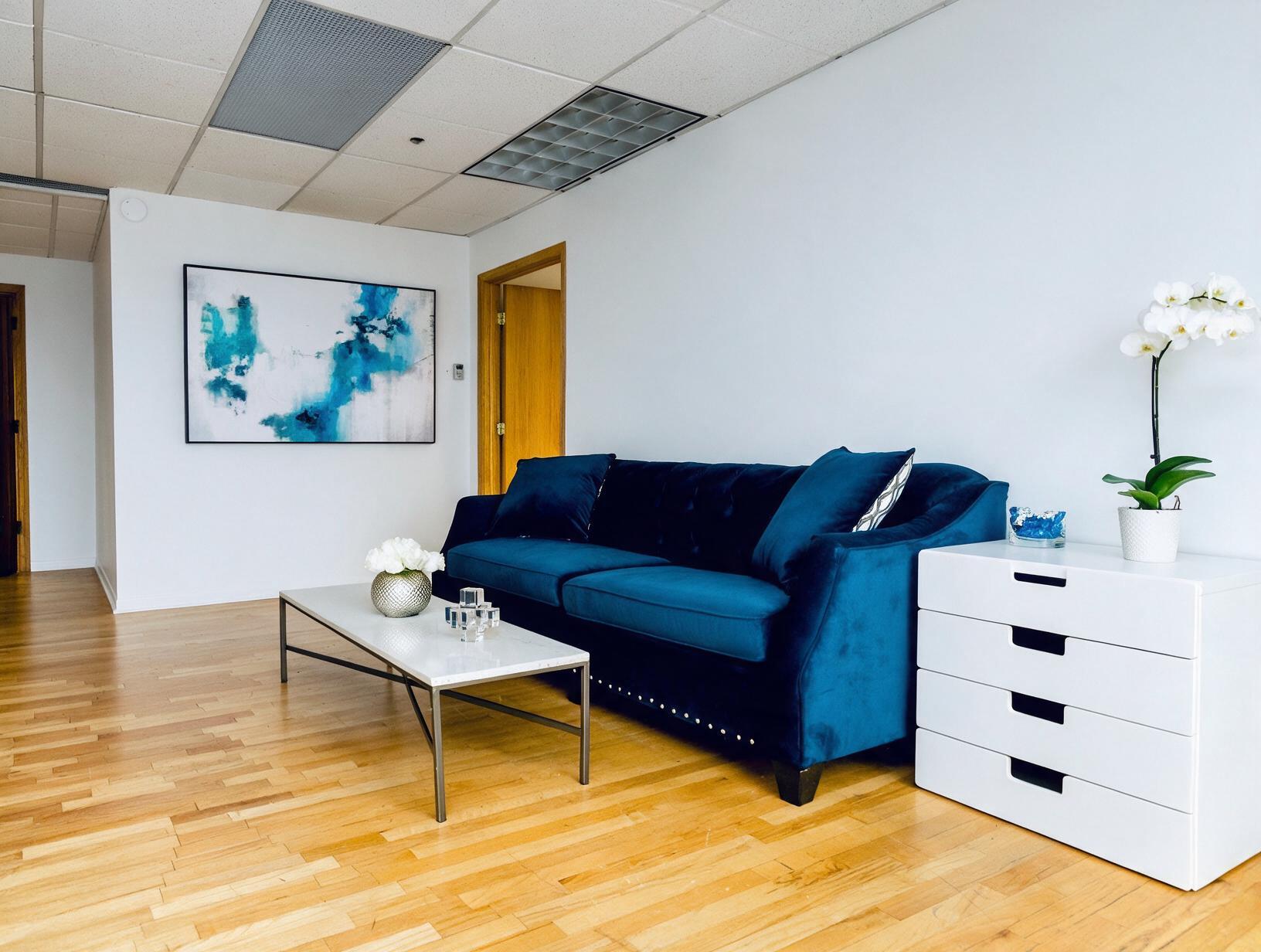 Tidy room with a blue velvet sofa, marble coffee table, white drawer unit with an orchid, abstract painting and wood floor.
