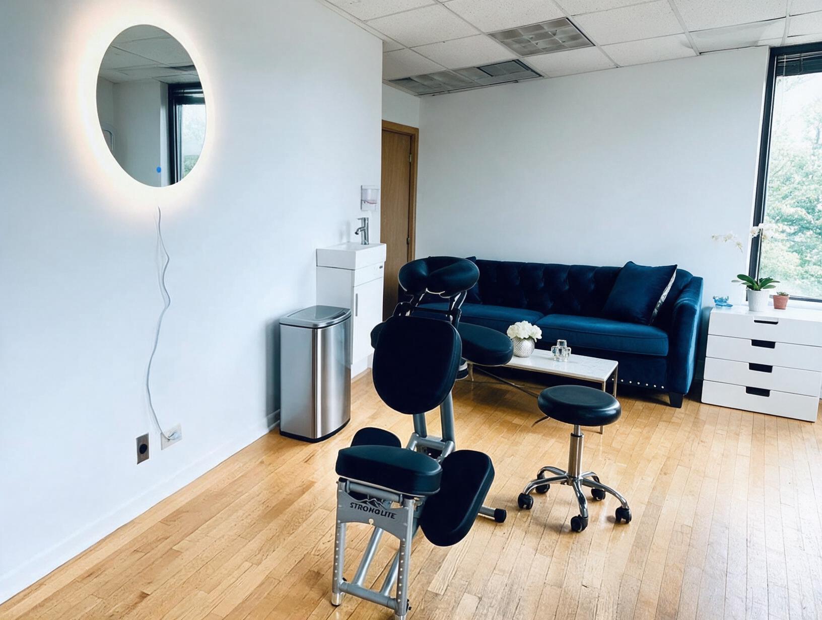 Bright empty treatment room with a black portable massage chair, round stool, blue velvet sofa